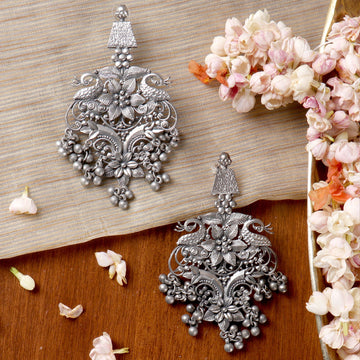 Intricately designed German silver earrings with floral and peacock motifs on wooden surface with flowers, German Silver Earrings Collection