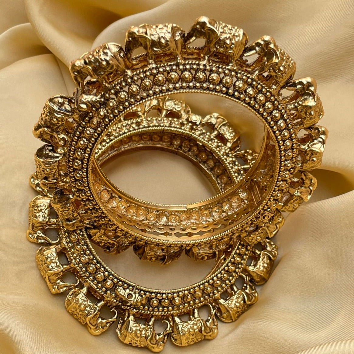 Intricately designed gold bangles featuring elephant motifs and detailed beadwork on a smooth fabric background