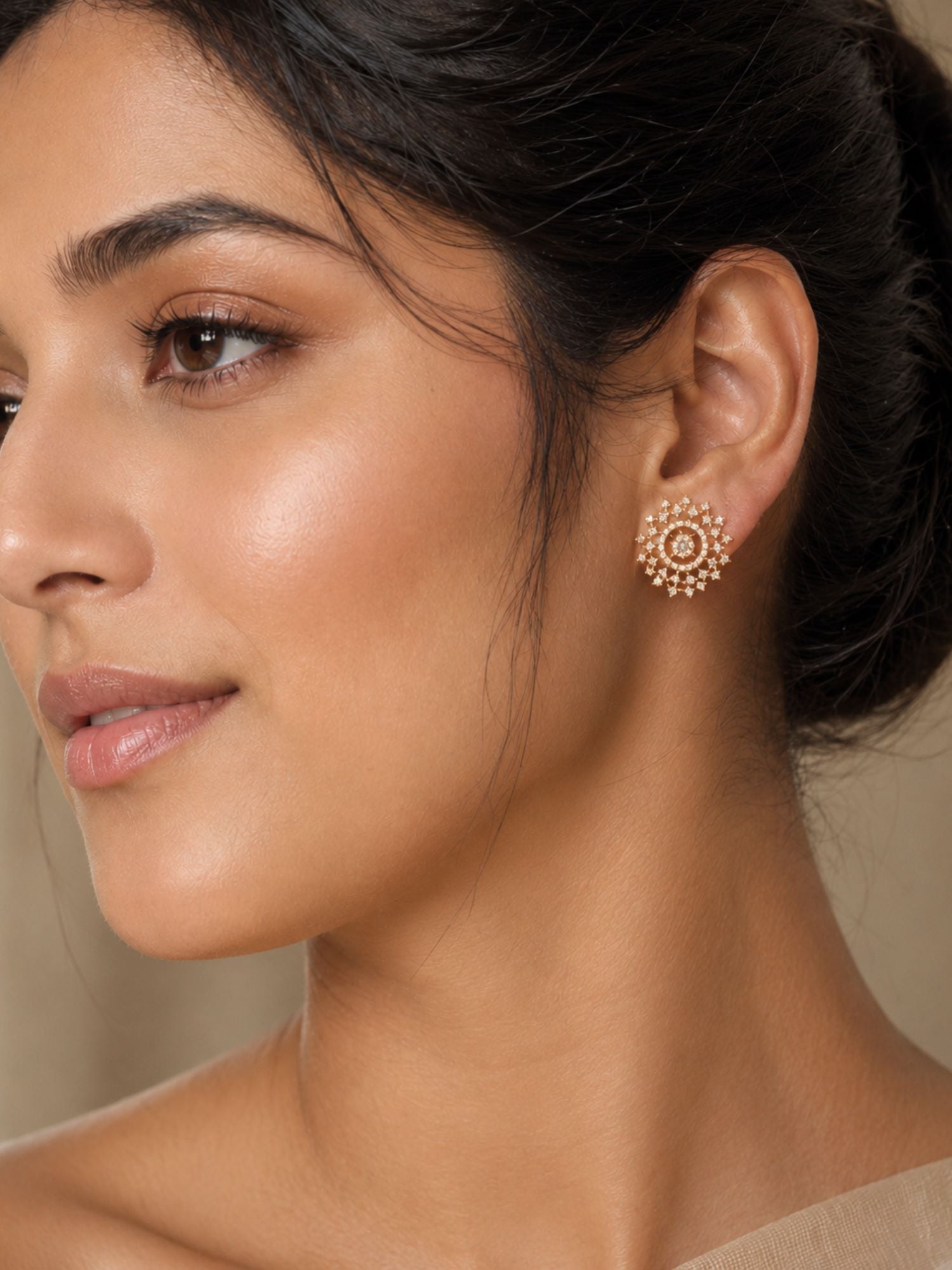 A close-up of a woman's ear wearing a small gold-toned mandarin-style stud earring against a soft beige background.