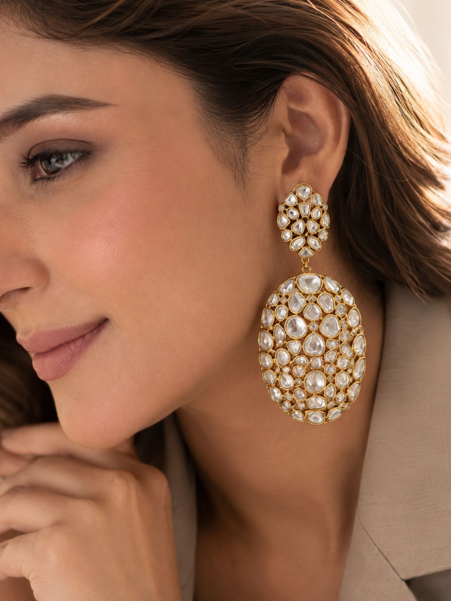 A close-up lifestyle shot of a woman wearing a large gold-plated drop earring adorned with clear crystals and white pearls. The earring features a round cluster of crystals at the top and a larger oval drop with a dense pavé pattern of white pearls and clear stones.