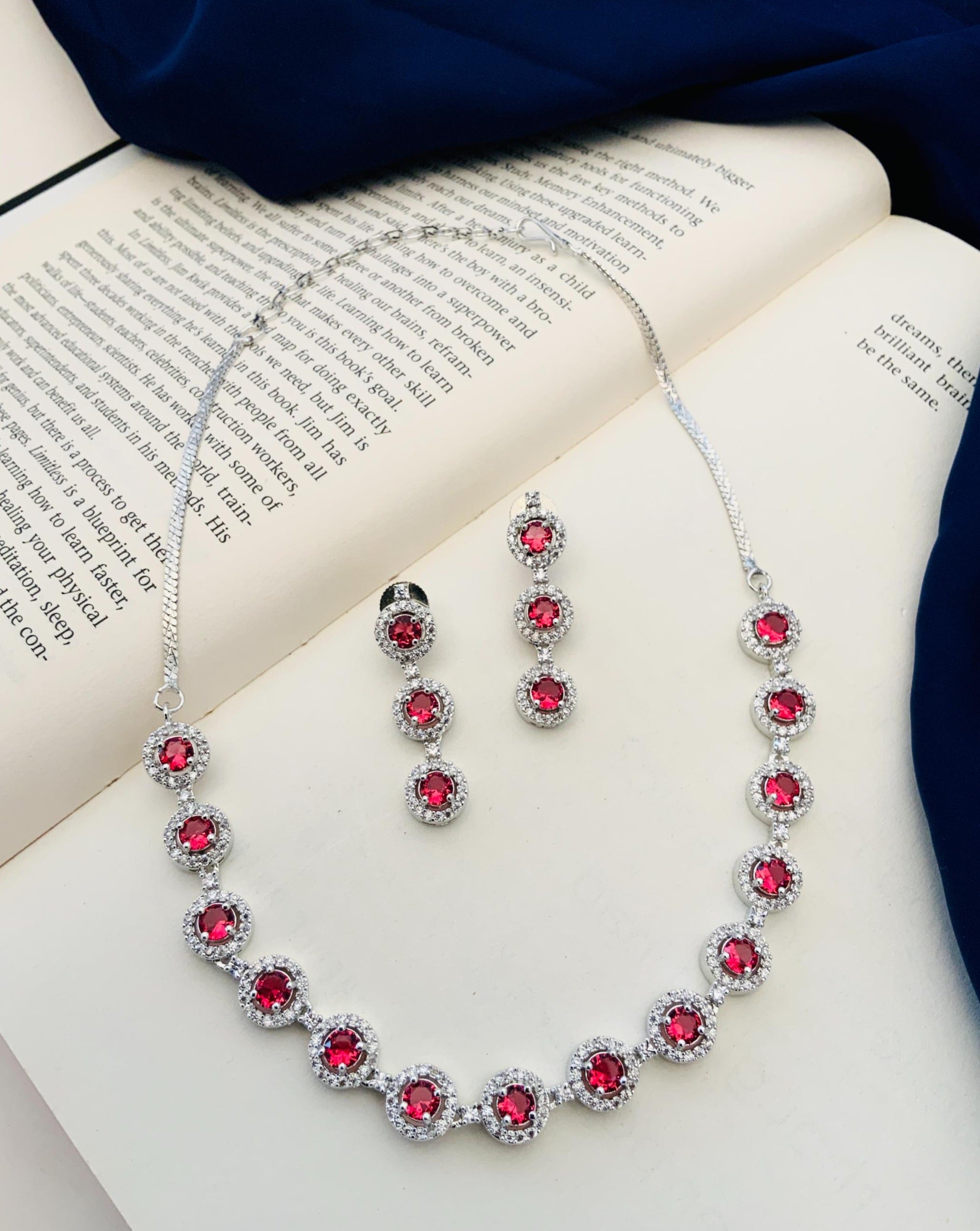 Abdesigns Silver Plated American Diamond Necklace Set with red round stones and matching drop earrings on open book background