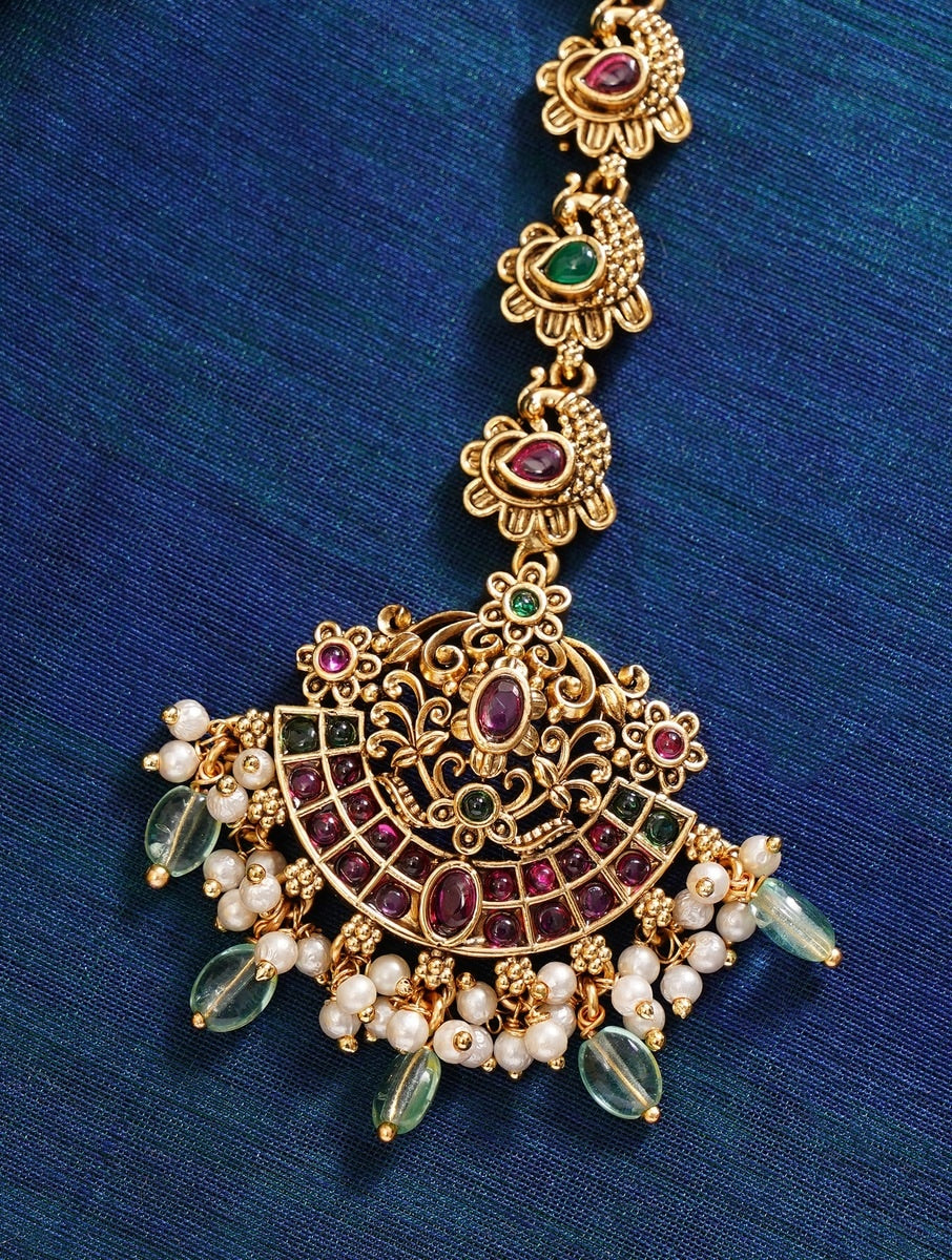 Matt Gold Plated KempStone Studed Maangtikka with intricate design and pearl accents on blue fabric background