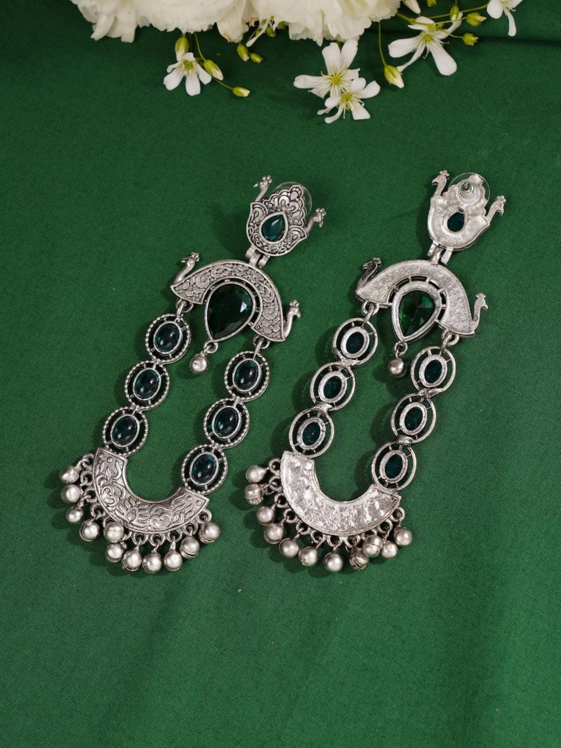 Long Peacock German Silver Earrings by Abdesignsjewellery with intricate detailing and dangling bells on green background